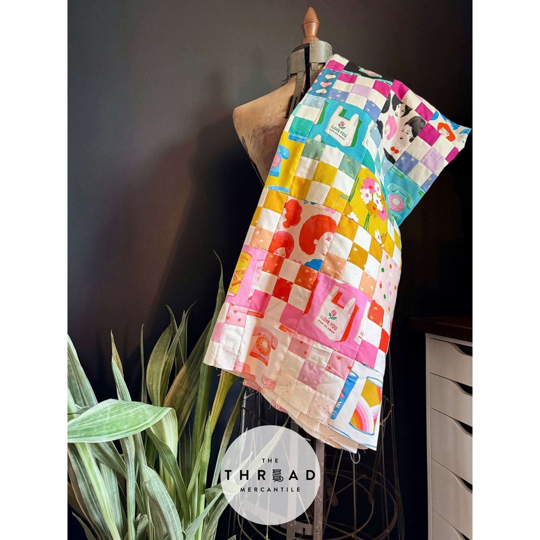 Vibrantly colored modern quilt draped over an antique mannequin in a modern setting; black wall & snake plant surround the mannequin.